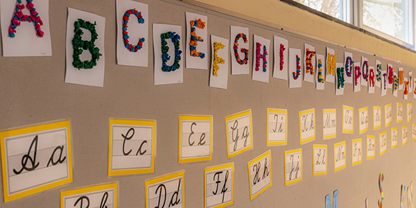 A classroom bulletin board displays alphabet posters featuring both colorful, textured block letters and traditional cursive script cards.
